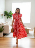 Swanky B Boutique red floral print midi dress with flutter sleeves, V-neck, and tie waist. Model is wearing heels and carrying a straw bag.